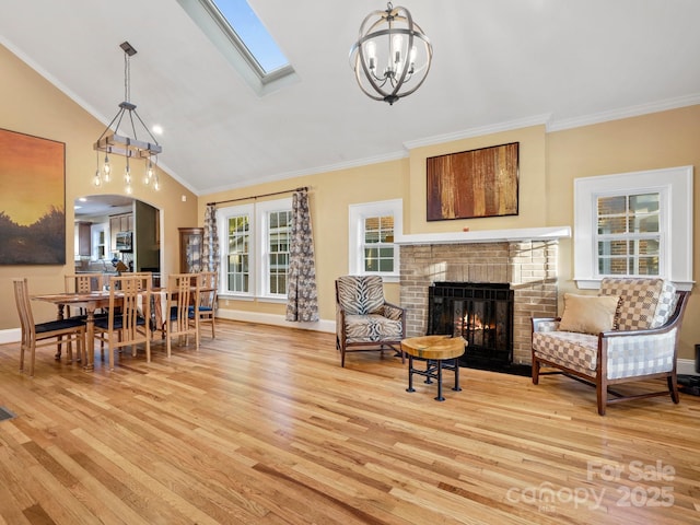 living room featuring vaulted ceiling with skylight, ornamental molding, light hardwood / wood-style floors, a brick fireplace, and an inviting chandelier