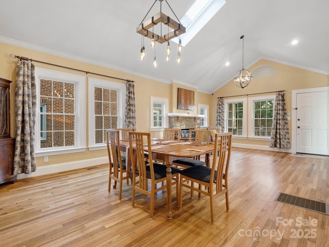 dining area featuring a stone fireplace, a chandelier, light wood-type flooring, ornamental molding, and vaulted ceiling with skylight