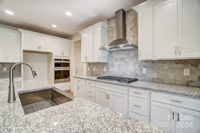 kitchen with wall chimney range hood, sink, light stone countertops, white cabinets, and appliances with stainless steel finishes