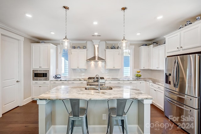 kitchen featuring an island with sink, white cabinets, wall chimney range hood, hanging light fixtures, and appliances with stainless steel finishes