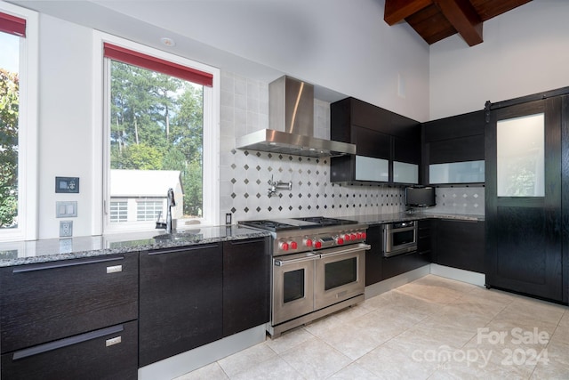 kitchen featuring light tile patterned flooring, range with two ovens, tasteful backsplash, wall chimney range hood, and dark cabinets