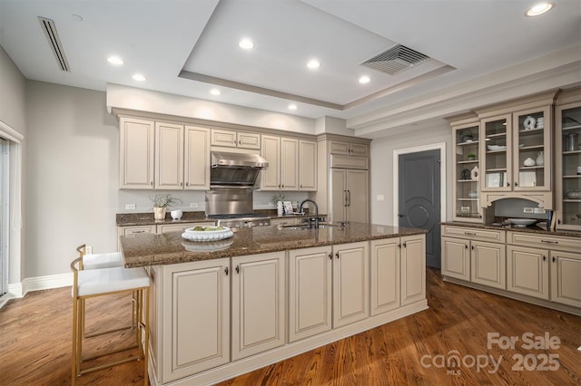 kitchen with sink, dark hardwood / wood-style flooring, dark stone counters, paneled built in fridge, and a center island with sink