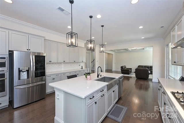 kitchen featuring dark wood-type flooring, a center island with sink, crown molding, decorative light fixtures, and appliances with stainless steel finishes