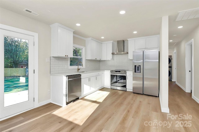 kitchen with sink, stacked washing maching and dryer, stainless steel appliances, white cabinets, and wall chimney exhaust hood