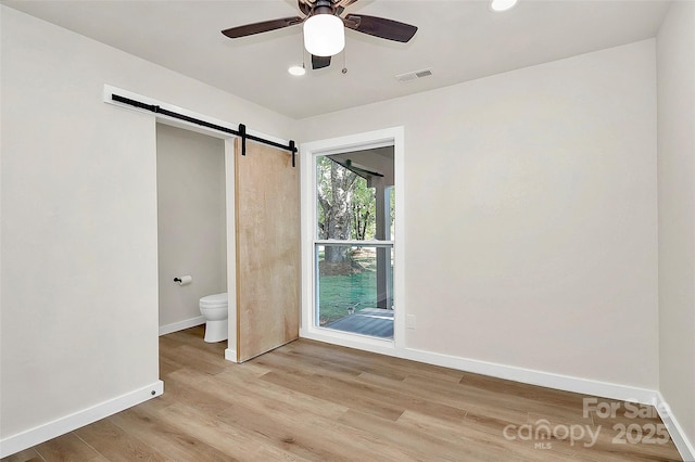 unfurnished bedroom with ensuite bathroom, a barn door, ceiling fan, and light hardwood / wood-style flooring