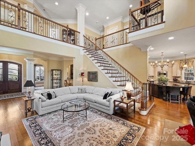 living room featuring a notable chandelier, wood-type flooring, a towering ceiling, ornamental molding, and ornate columns