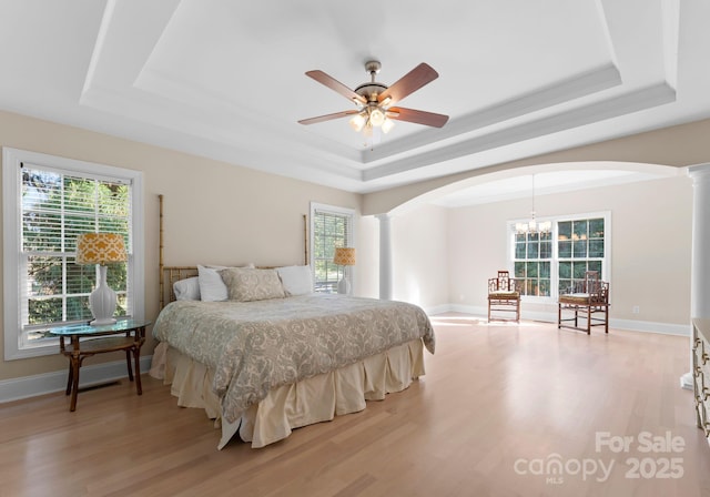 bedroom with a tray ceiling, ornate columns, ceiling fan with notable chandelier, and light wood-type flooring