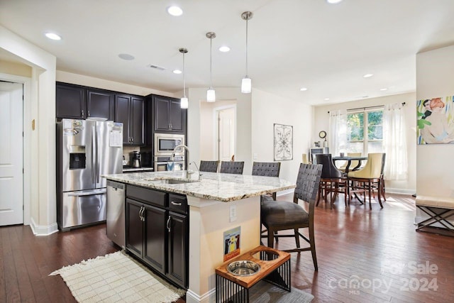 kitchen with a kitchen island with sink, dark wood-type flooring, sink, appliances with stainless steel finishes, and decorative light fixtures