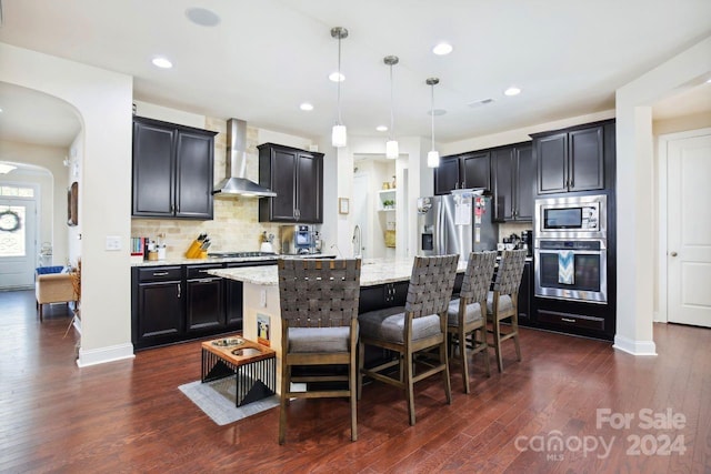 kitchen featuring wall chimney exhaust hood, dark wood-type flooring, light stone counters, hanging light fixtures, and appliances with stainless steel finishes