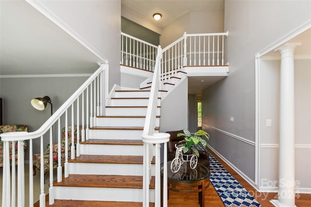 stairway with a towering ceiling, wood-type flooring, crown molding, and decorative columns