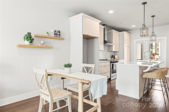 kitchen with an island with sink, dark wood-type flooring, wall chimney range hood, hanging light fixtures, and stainless steel range