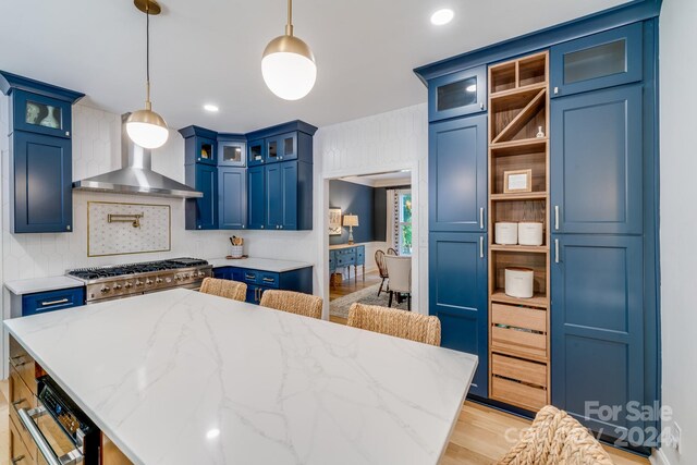 kitchen with wall chimney range hood, blue cabinetry, light hardwood / wood-style flooring, light stone counters, and hanging light fixtures