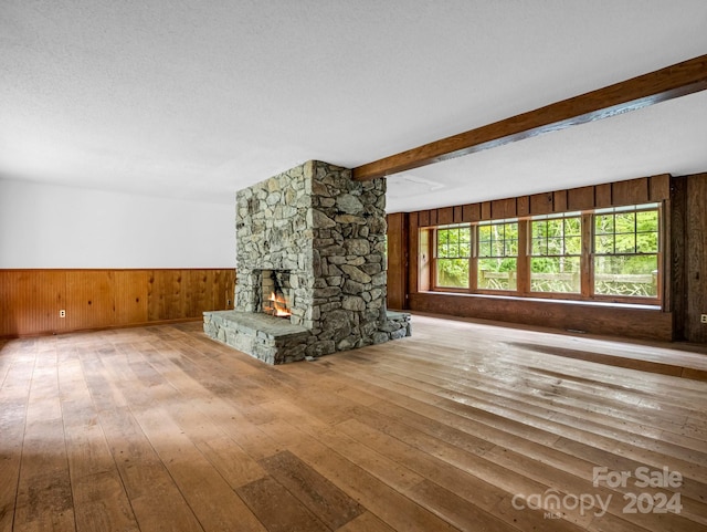 unfurnished living room with wood walls, light hardwood / wood-style floors, beam ceiling, a stone fireplace, and a textured ceiling