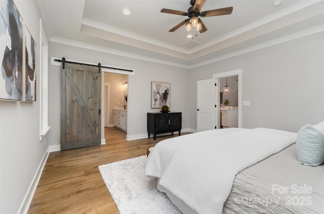 bedroom featuring a barn door, light wood-style flooring, a tray ceiling, ornamental molding, and ensuite bath