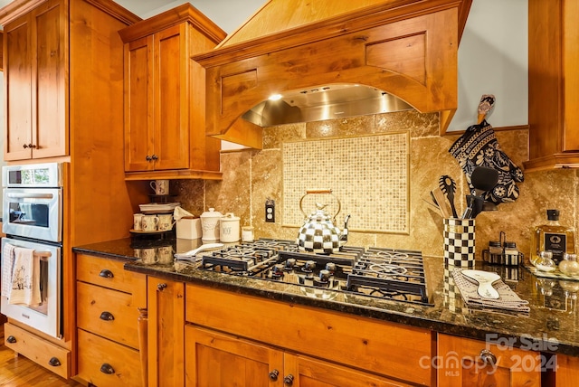 kitchen featuring gas stovetop, decorative backsplash, wall chimney range hood, wood-type flooring, and dark stone countertops