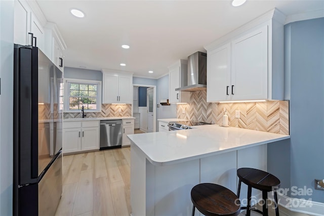 kitchen featuring kitchen peninsula, wall chimney exhaust hood, white cabinetry, stainless steel appliances, and a breakfast bar