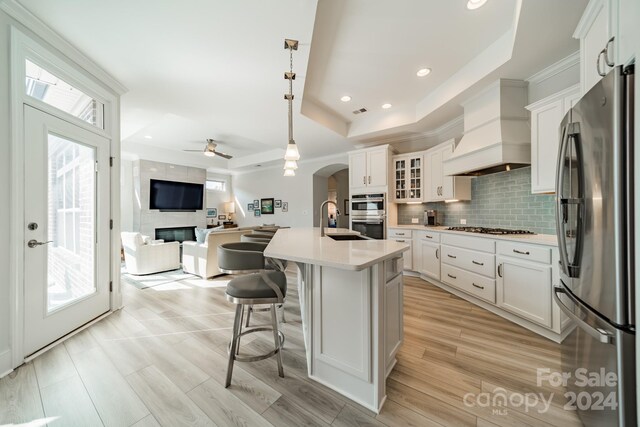 kitchen featuring stainless steel appliances, a tray ceiling, a center island with sink, decorative light fixtures, and white cabinetry