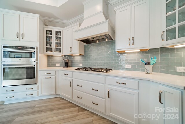 kitchen with decorative backsplash, white cabinetry, light hardwood / wood-style flooring, custom range hood, and stainless steel appliances