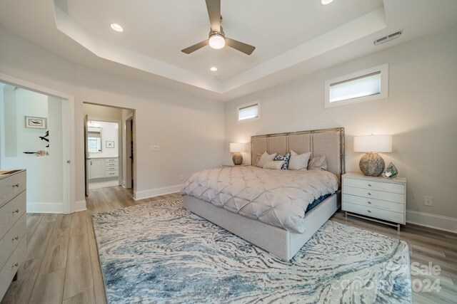 bedroom with ensuite bath, a raised ceiling, light wood-type flooring, and ceiling fan