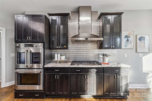 kitchen featuring appliances with stainless steel finishes, decorative backsplash, hardwood / wood-style flooring, light stone countertops, and wall chimney range hood