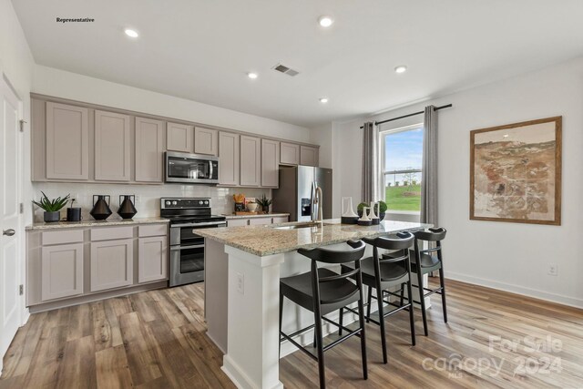 kitchen featuring stainless steel appliances, light stone counters, an island with sink, gray cabinets, and light wood-type flooring