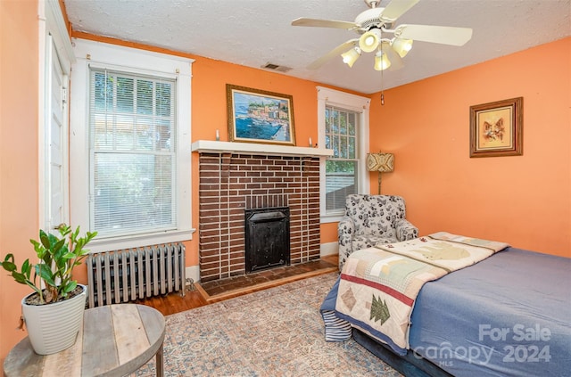 bedroom with a textured ceiling, radiator heating unit, a brick fireplace, hardwood / wood-style floors, and ceiling fan