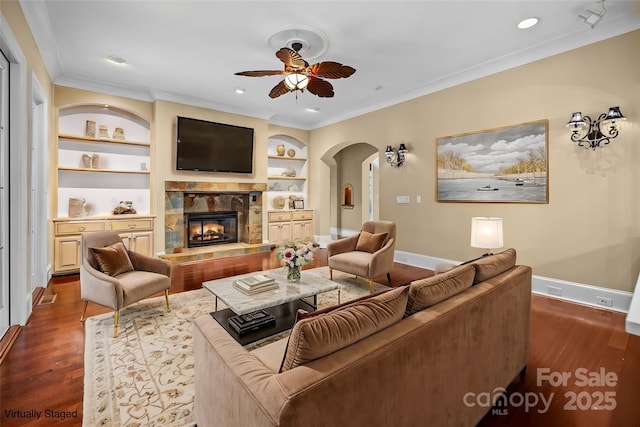 living room with built in shelves, dark wood-type flooring, a fireplace, baseboards, and crown molding