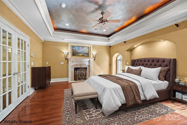 bedroom featuring wood finished floors, a fireplace with flush hearth, ornamental molding, french doors, and a tray ceiling