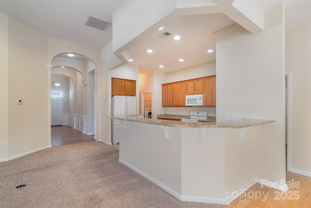 kitchen featuring a breakfast bar, white appliances, light stone counters, light colored carpet, and kitchen peninsula