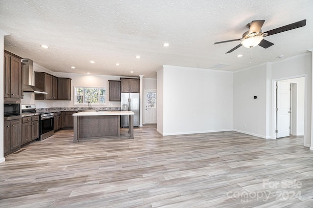 kitchen featuring wall chimney range hood, light hardwood / wood-style flooring, a textured ceiling, a kitchen island, and appliances with stainless steel finishes