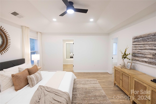 bedroom with a tray ceiling, ensuite bathroom, light hardwood / wood-style floors, and ceiling fan