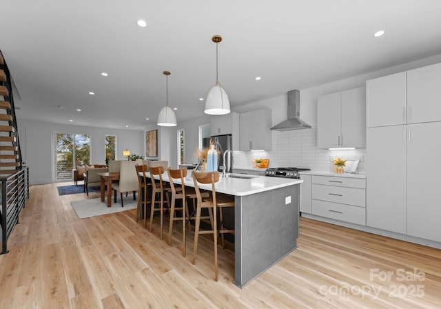 kitchen with decorative light fixtures, an island with sink, sink, white cabinets, and wall chimney exhaust hood