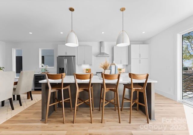 kitchen featuring stainless steel fridge with ice dispenser, an island with sink, hanging light fixtures, and wall chimney exhaust hood