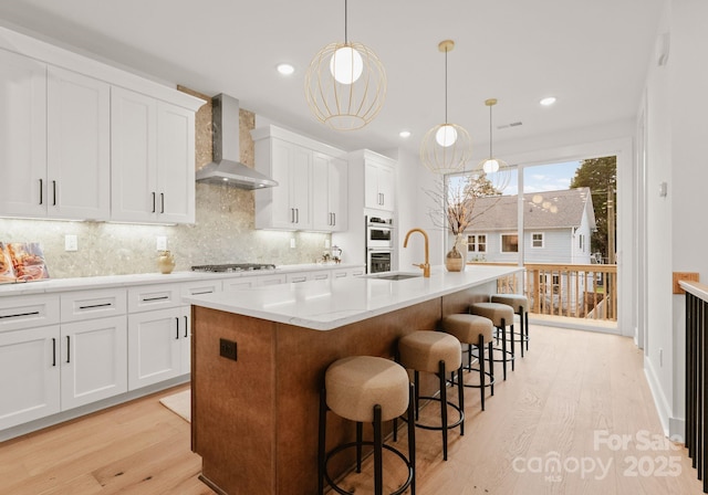 kitchen featuring a center island with sink, white cabinets, hanging light fixtures, and wall chimney range hood