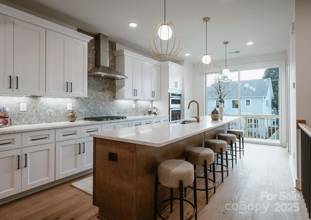 kitchen with hanging light fixtures, white cabinetry, tasteful backsplash, wall chimney range hood, and a center island with sink