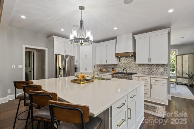 kitchen featuring stainless steel refrigerator with ice dispenser, white cabinetry, an island with sink, premium range hood, and sink