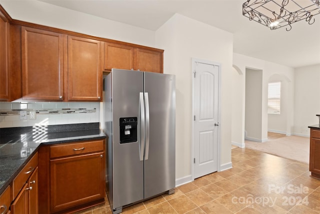 kitchen with an inviting chandelier, dark stone countertops, stainless steel fridge, decorative backsplash, and light tile patterned floors