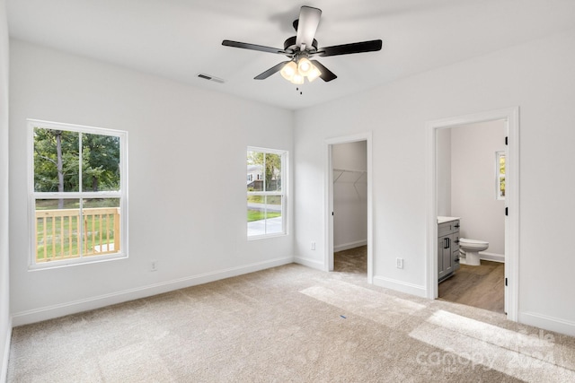 unfurnished bedroom with ensuite bathroom, a walk in closet, a closet, light carpet, and ceiling fan