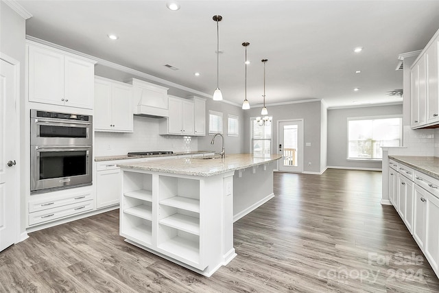 kitchen featuring white cabinets, hanging light fixtures, stainless steel double oven, and a kitchen island with sink