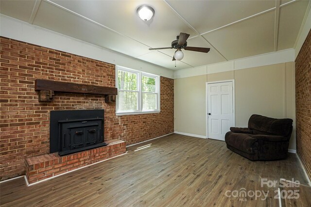 unfurnished living room with wood finished floors, ceiling fan, brick wall, baseboards, and a wood stove