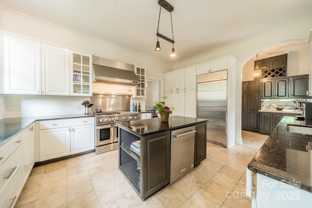 kitchen with arched walkways, high end appliances, wall chimney range hood, white cabinetry, and backsplash