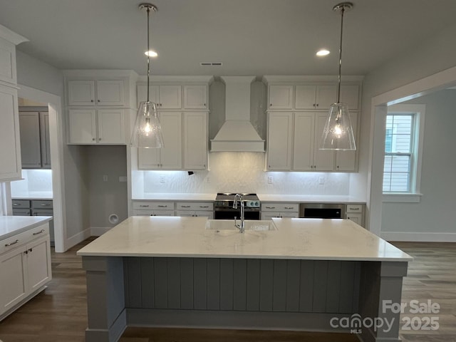 kitchen featuring premium range hood, decorative light fixtures, an island with sink, and light stone counters