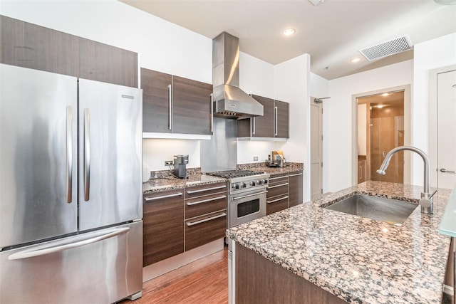 kitchen with light stone countertops, appliances with stainless steel finishes, sink, wall chimney exhaust hood, and light hardwood / wood-style flooring