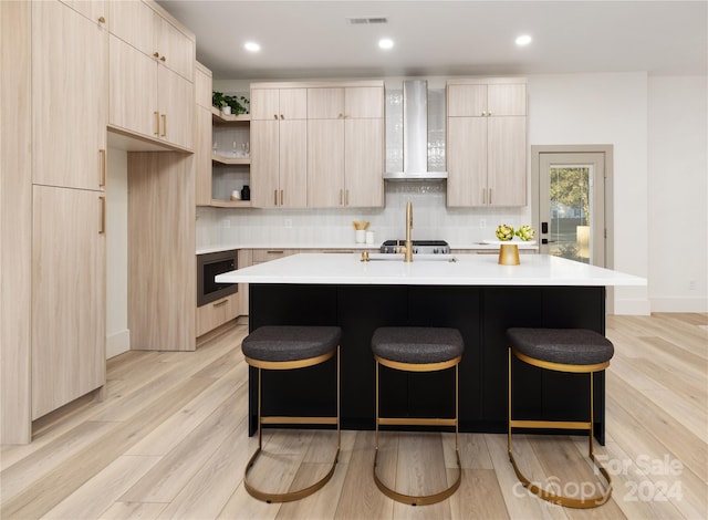 kitchen with a kitchen breakfast bar, a kitchen island with sink, wall chimney range hood, light brown cabinets, and light hardwood / wood-style floors