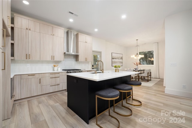 kitchen with light brown cabinetry, wall chimney exhaust hood, an inviting chandelier, light hardwood / wood-style floors, and an island with sink