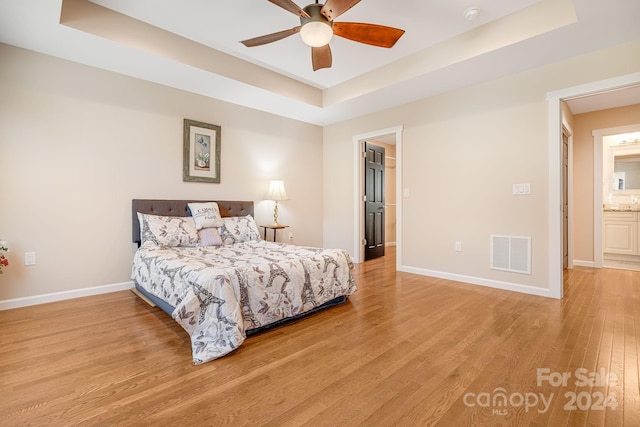 bedroom with ceiling fan, a raised ceiling, light wood-type flooring, and connected bathroom