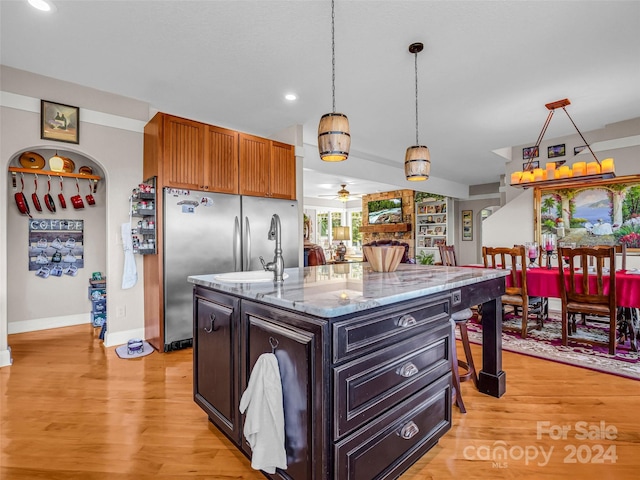 kitchen with stainless steel fridge, light hardwood / wood-style floors, ceiling fan, light stone counters, and a center island with sink