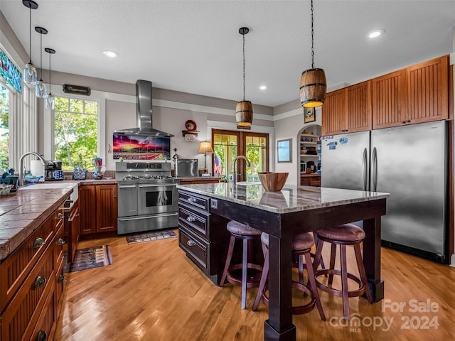 kitchen with wall chimney range hood, a kitchen breakfast bar, a center island with sink, appliances with stainless steel finishes, and light hardwood / wood-style floors