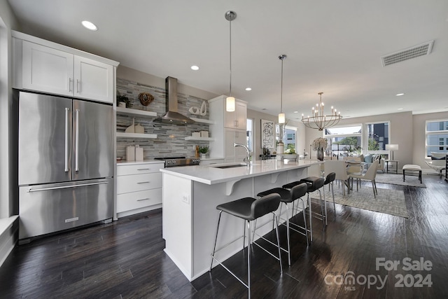 kitchen featuring wall chimney exhaust hood, an island with sink, stainless steel appliances, pendant lighting, and white cabinetry