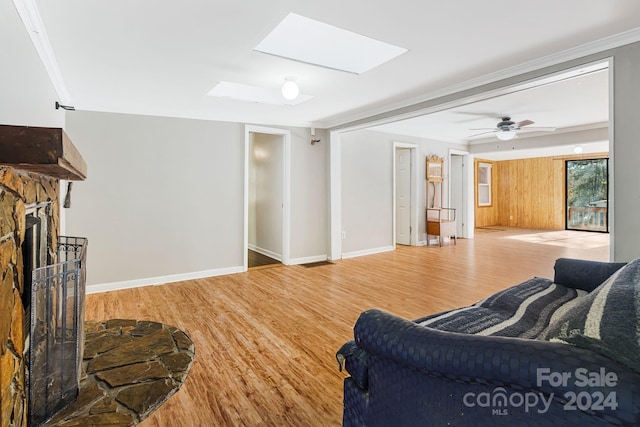 living room featuring a stone fireplace, light hardwood / wood-style flooring, ornamental molding, a skylight, and ceiling fan